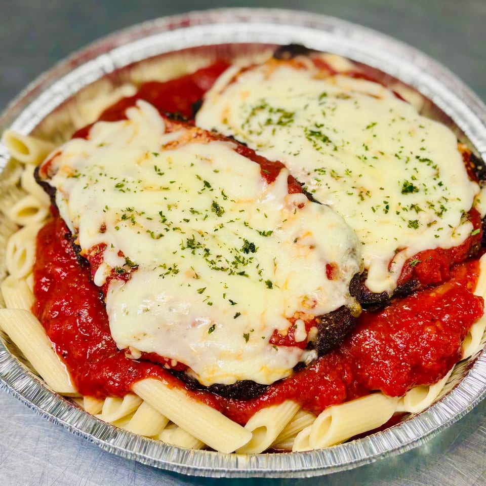 Baked eggplant Parmesan topped with melted mozzarella and herbs, served over penne pasta in a foil tray.
